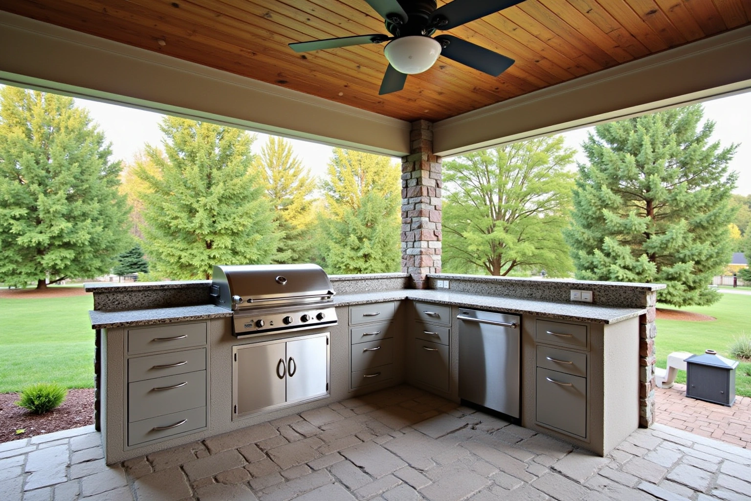Covered patio outdoor kitchen with grey drawers, stainless access doors, a built-in grill and granite counters, trees beyond.